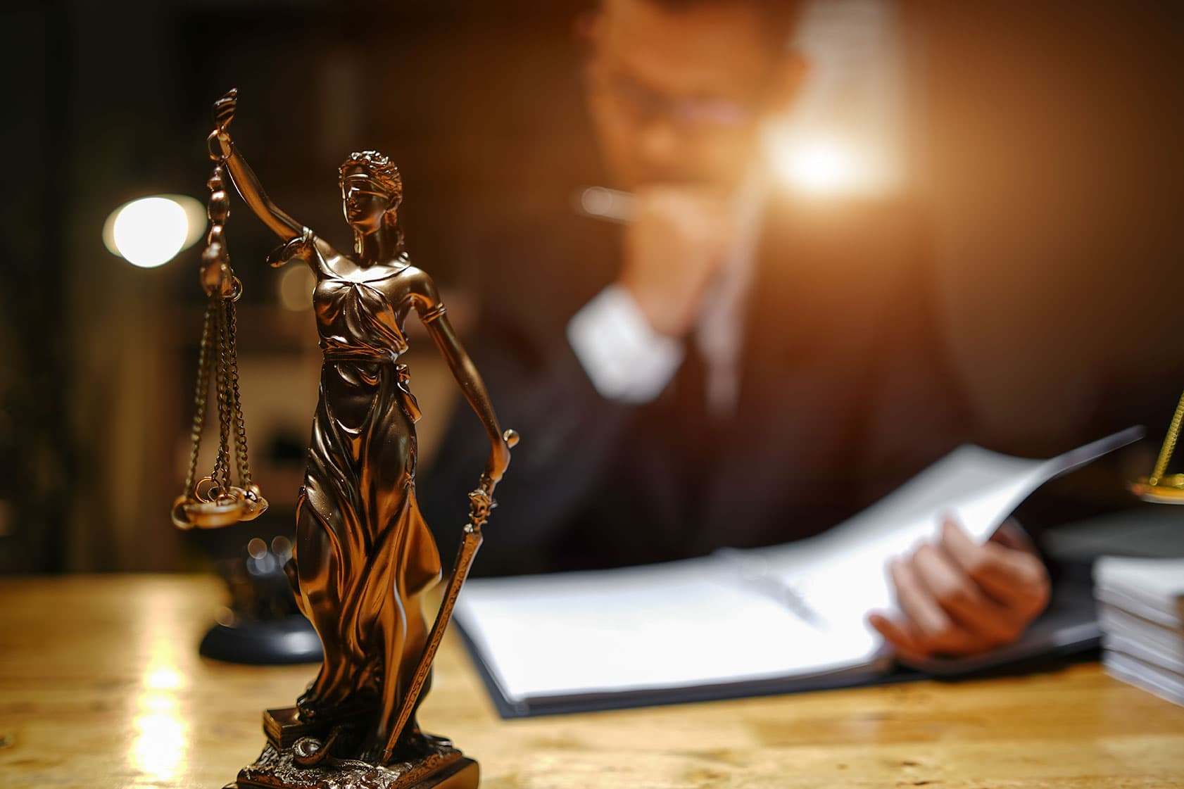 Gold Lady Justice statue symbolizing law and fairness, with a blurred lawyer researching legal documents in background, representing legal expertise and justice advocacy.